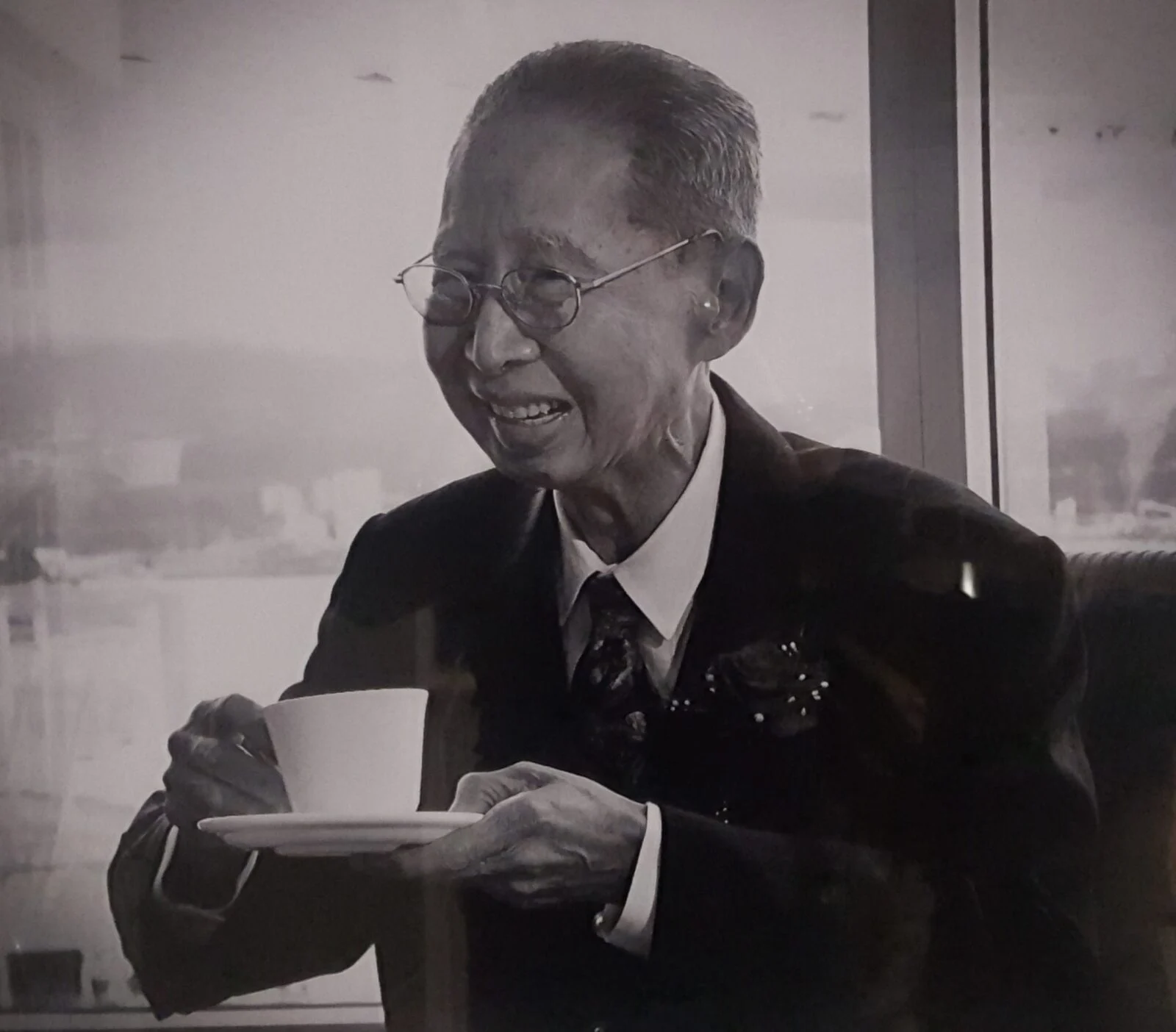 Homme âgé souriant en costume tenant une tasse et une soucoupe dans un cadre intérieur lumineux