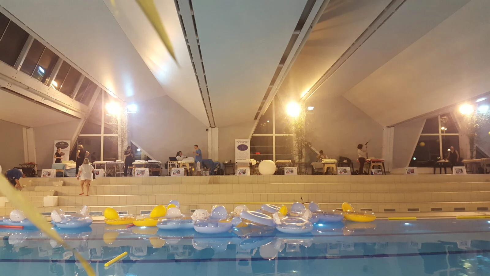 Bassin de piscine intérieur avec bouées jaunes et bleues flottantes, tables de massage et personnes en arrière-plan