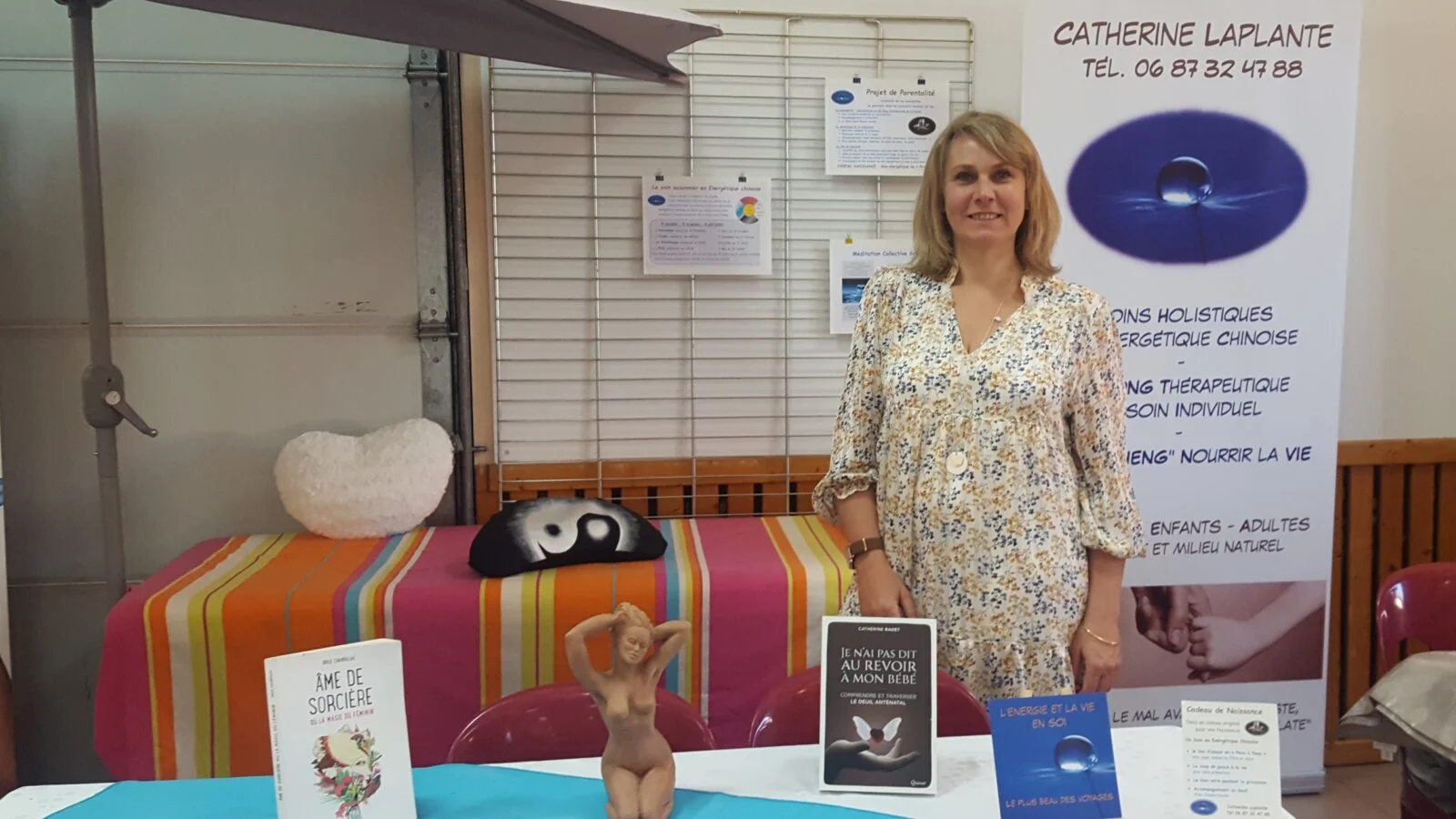 Femme debout devant un stand de livres de bien-être avec des affiches et une bannière, salon de soins holistiques.