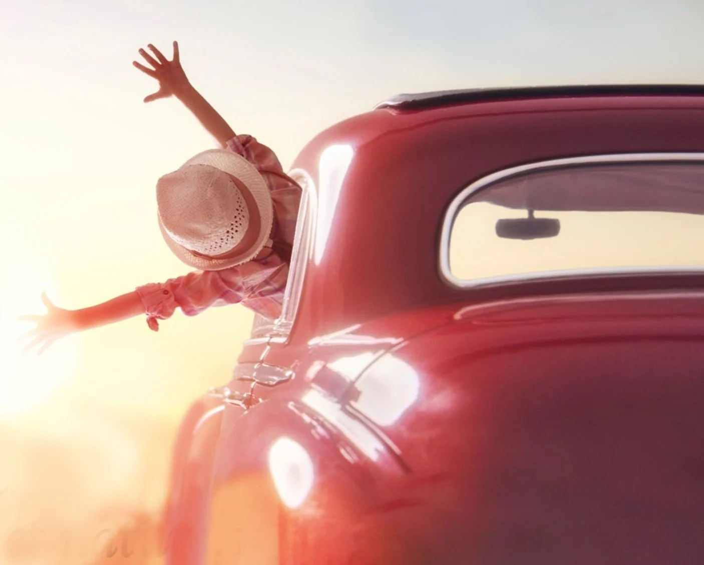 Enfant avec chapeau, bras étendus vers le soleil, sortant d'une voiture rouge.