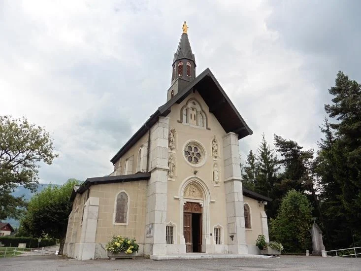 Église en pierre entourée d'arbres sous un ciel nuageux.