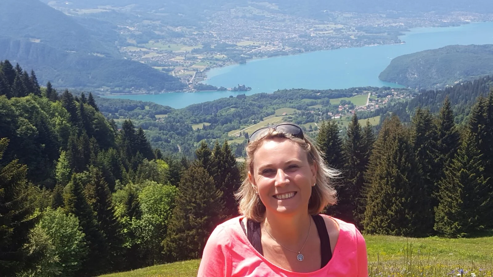 Femme souriante devant un paysage alpin avec lac et forêts.