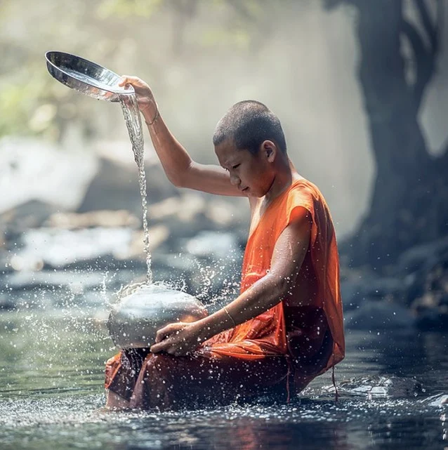 Jeune moine bouddhiste en robe orange versant de l'eau dans un ruisseau.