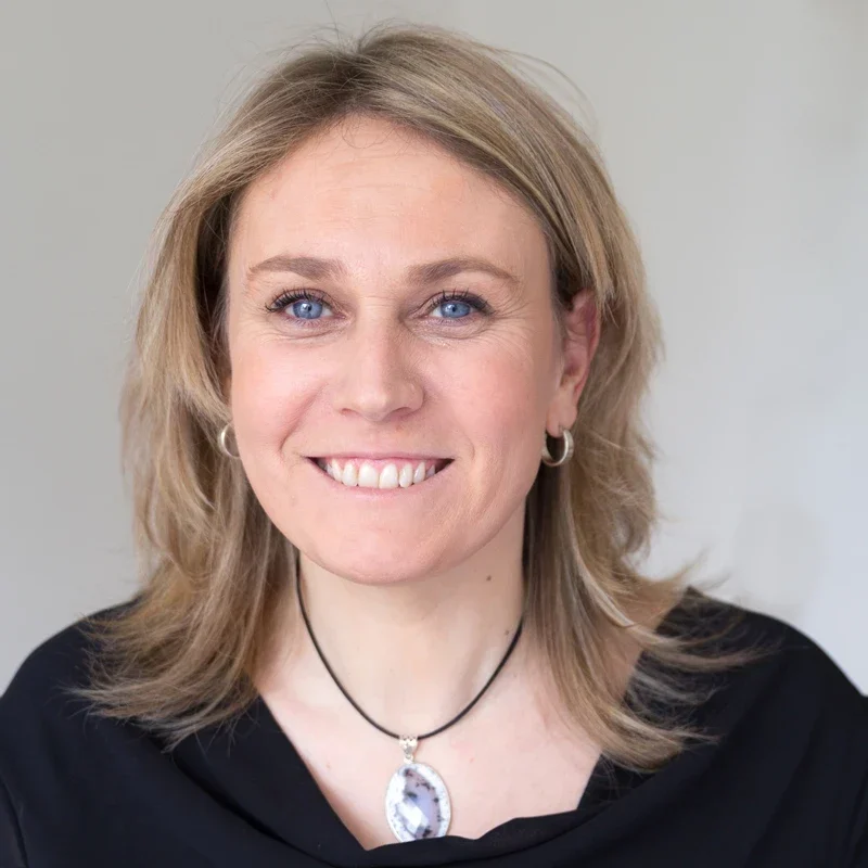 Femme souriante aux cheveux blonds portant un collier et une blouse noire.